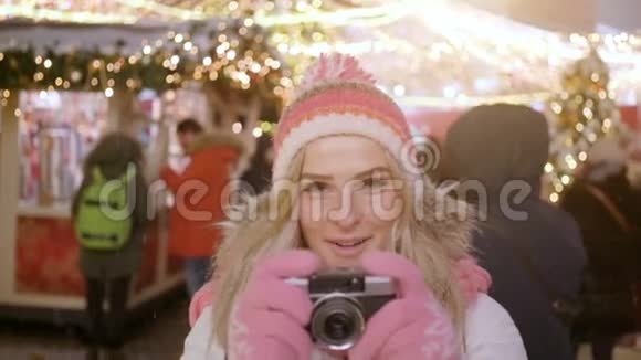 年轻的金发女郎微笑着穿着温暖的衣服用复古相机在冬季市场的背景上拍摄一张照片视频的预览图