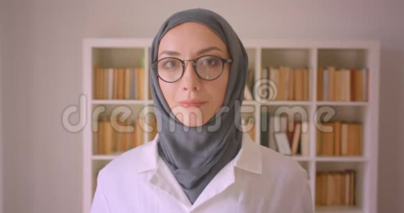 一幅年轻的穆斯林女医生戴着眼镜戴着头巾在室内图书馆里看着相机开心地微笑的特写照片视频的预览图