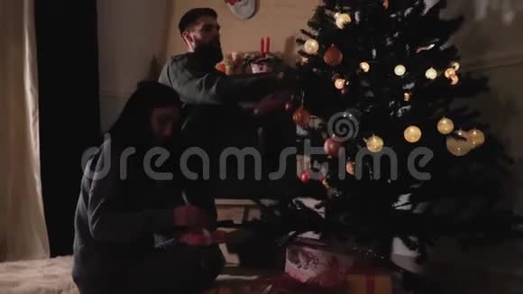 恋爱中的男女在家里装饰圣诞树女人纠正礼品盒上的蝴蝶结幸福夫妻准备视频的预览图