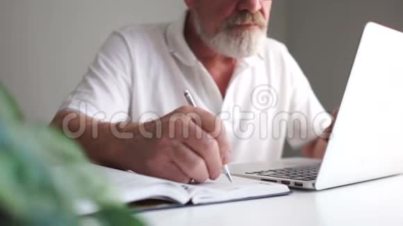 一个留着眼镜和一件白色T恤的胡子的男人在笔记本上做笔记并使用笔记本电脑视频的预览图