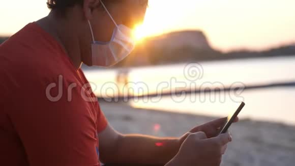 COVID19男人戴着面具坐在海滩上在日落时使用手机冠状病毒流行视频的预览图