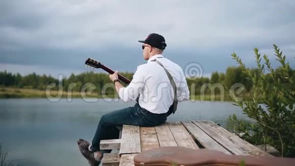 酷吉他手坐在湖边的码头上演奏在运动的电影视频的预览图