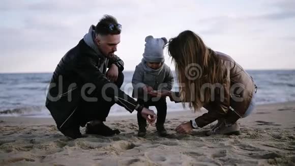 爸爸妈妈带着孩子蹒跚学步的小男孩在寒冷多云多风的天气里玩沙子男人帮助小男孩视频的预览图