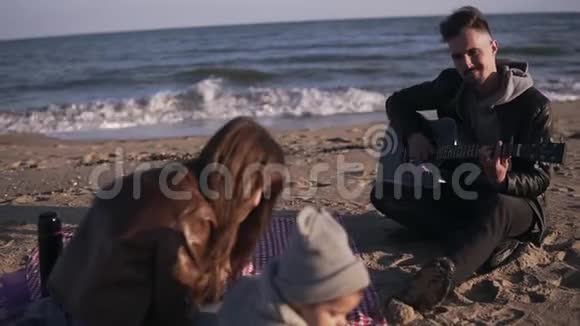 一个快乐微笑的家庭坐在沙滩上的格子毯子上父亲和母亲一起弹吉他视频的预览图