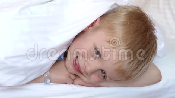 一个可爱的金发男孩躺在白色毯子下的床上头上盖着一条毯子视频的预览图