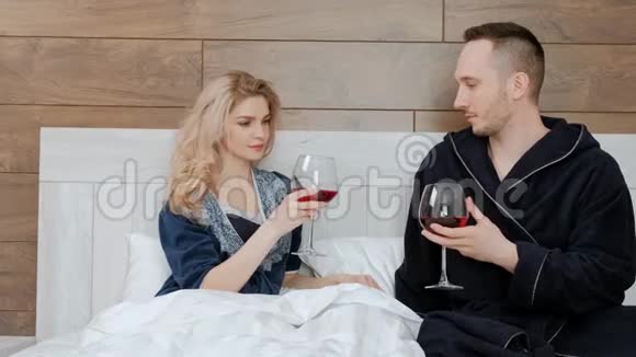 年轻的已婚夫妇穿着浴袍喝着红酒躺在酒店房间的卧室里的一张白床上视频的预览图