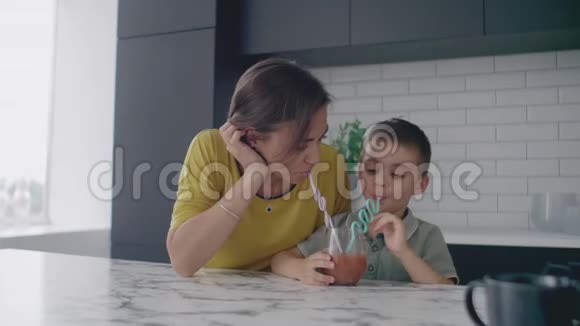 爱美丽的母亲和儿子一起微笑着从一个玻璃杯中通过一个管子喝橙汁视频的预览图