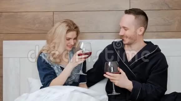 年轻的已婚夫妇穿着浴袍喝着红酒躺在酒店房间的卧室里的一张白床上视频的预览图