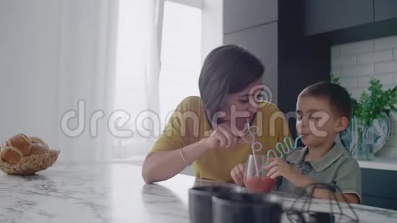 爱美丽的母亲和儿子一起微笑着从一个玻璃杯中通过一个管子喝橙汁视频的预览图