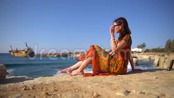 可爱的年轻女子穿着夏装戴着太阳镜坐在岩石海岸边以一幅古老的风景画为背景视频的预览图