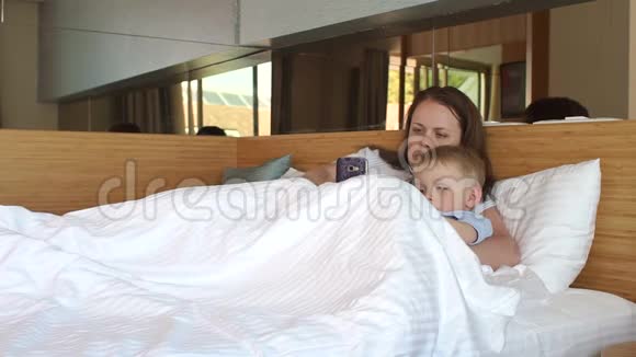 妈妈和儿子躺在毯子下面的床上在电话里玩游戏视频的预览图