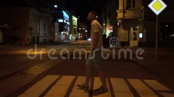 穿着条纹马球和绿色短裤的长胡子男人晚上在人行横道上穿过马路视频的预览图