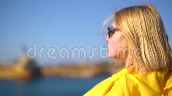 一个金发女孩的特写镜头戴着太阳镜和一件黄色雨衣站在阳光明媚的大风天气下的悬崖上视频的预览图