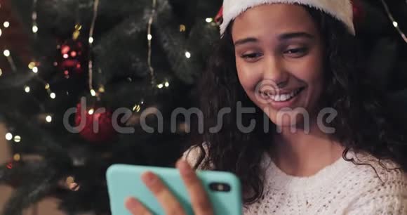 圣诞老人帽中快乐美丽的穆拉托女孩坐在圣诞树的背景下用她的智能手机看着视频的预览图