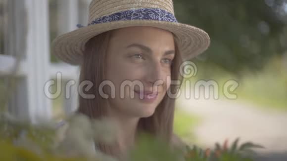 一个妩媚的年轻女子戴着草帽穿着白色连衣裙坐在小村屋前农村视频的预览图