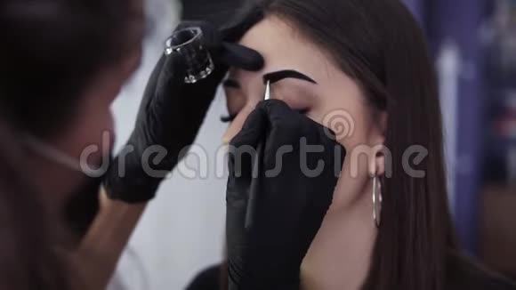 审美者用棕色染料在年轻女子的眉毛上涂上深色油漆在一家美容工作室里进行着色特写视频的预览图