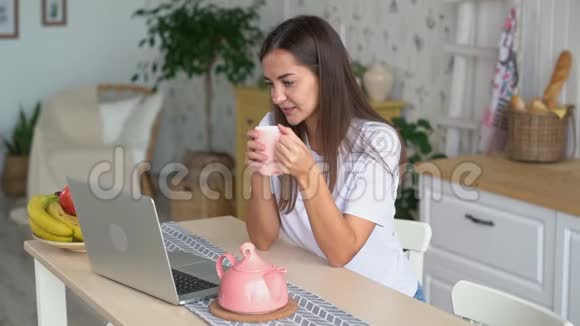 年轻女子坐在厨房里喝茶手提电脑看电影慢动作视频的预览图