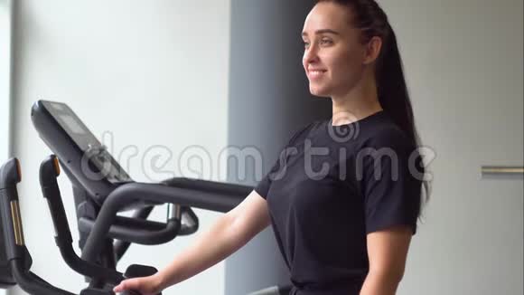 一个年轻漂亮的女孩骑着一辆静止的自行车在健身房里的肖像视频的预览图