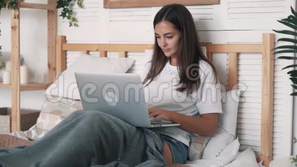 美丽的女孩坐在床上用笔记本电脑工作或在线聊天慢动作视频的预览图