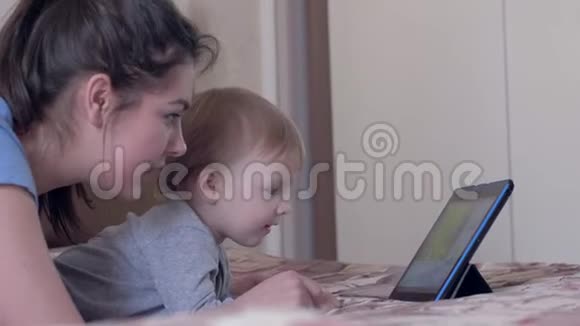 快乐的童年可爱的小男孩带着微笑的妈妈躺在家里的平板上看卡通片视频的预览图
