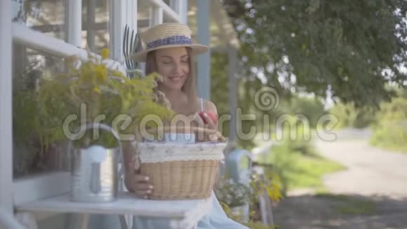美丽的年轻女子戴着草帽穿着白色连衣裙从小篮子里拿出苹果坐在小篮子前面视频的预览图