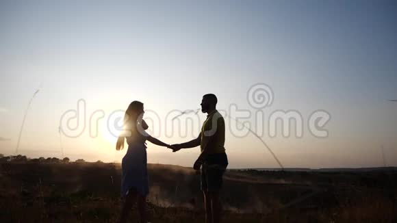 一对相爱的年轻夫妇的剪影在黄昏的夕阳灯光下与天空牵手农村的夫妇视频的预览图