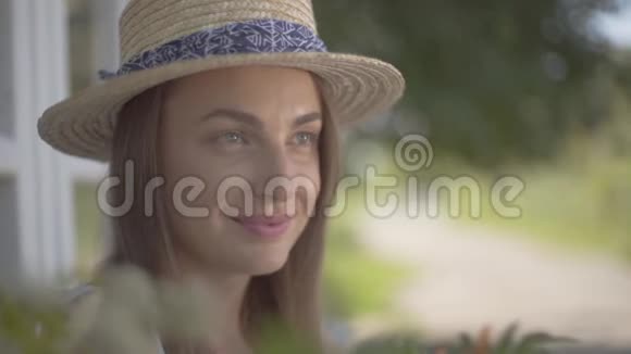 一个妩媚的年轻女子戴着草帽穿着白色连衣裙坐在小村屋前农村视频的预览图