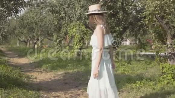 带着草帽和白色长裙的迷人年轻女子的后景穿过绿色的夏日花园视频的预览图