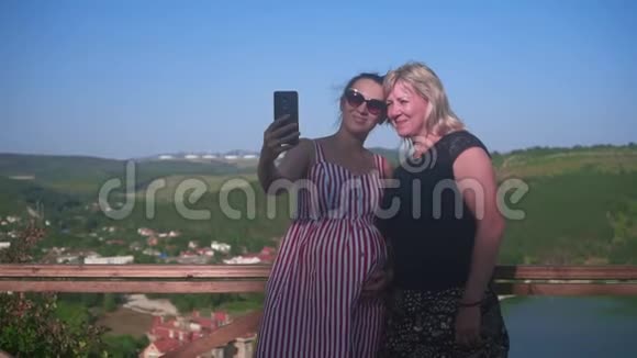 一个女人和一个怀孕的女孩正在一个山湖附近的高处行走女人喜欢在智能手机上自拍视频的预览图