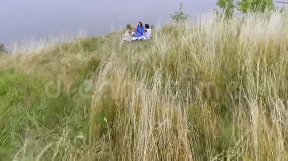 在湖边或河边坐着三个穿着夏日长礼服的女朋友的空中观景两个高加索人和一个视频的预览图