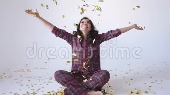 漂亮的快乐女人穿着睡衣坐在工作室里扔着金色的纸屑慢动作视频的预览图
