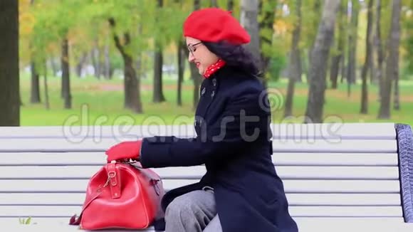 女人穿着红色的包在秋天公园的长凳上看东西教师节视频的预览图