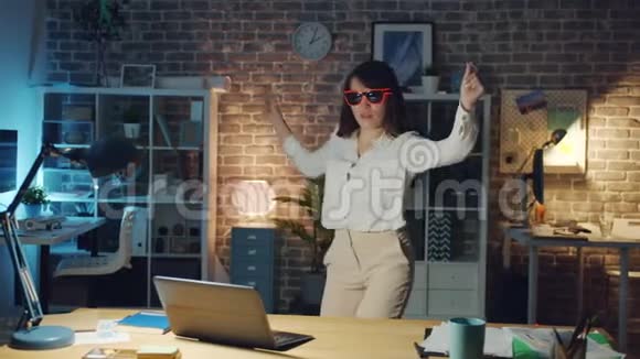 穿着时尚眼镜的快乐女孩在工作场所跳舞一个人在晚上玩视频的预览图