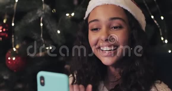 圣诞帽里美丽的微笑女孩坐在装饰好的圣诞树旁通过她的智能手机进行视频通话视频的预览图