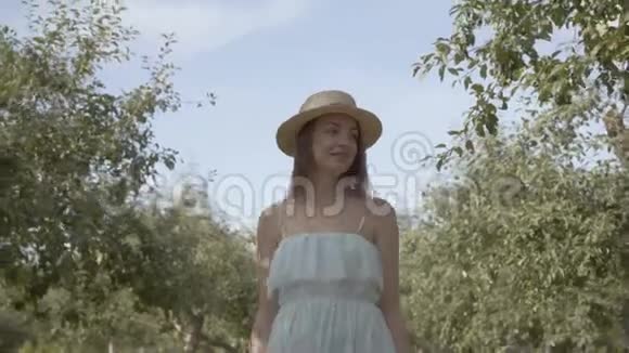 迷人的年轻微笑的女人戴着草帽穿着长长的白色连衣裙穿过绿色的夏天花园免费农村视频的预览图