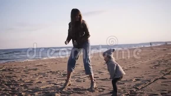 有趣的妈妈和穿着暖和的衣服的小宝宝在海边的沙滩上跳舞跳着跳着玩视频的预览图