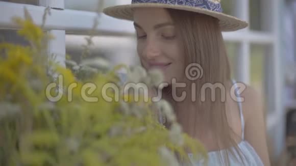 肖像美丽的年轻女子戴着草帽和白色的连衣裙坐在小村庄的房子前面农村视频的预览图