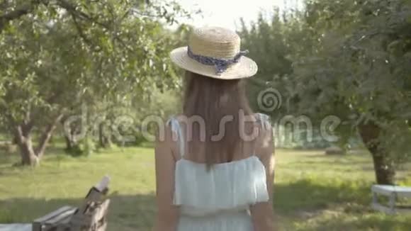 后景可爱的年轻女子戴着草帽和白色长裙走过绿色的夏天花园免费农村视频的预览图