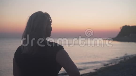 一个女人站在海上日落的背景上一个女人背对着镜头站在海滩上视频的预览图