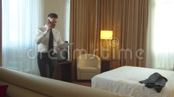 穿着衬衫和领带的人在酒店的房间里散步打电话和看文件视频的预览图