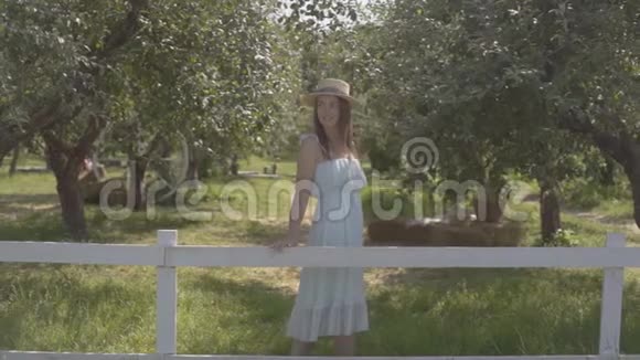 漂亮的年轻女子戴着草帽穿着白色长裙望着远处的镜头微笑着站在绿色的地方视频的预览图