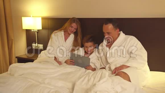 父母和小儿子坐在床上在酒店房间里看一些平板电脑视频的预览图