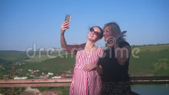一个女人和一个怀孕的女孩正在一个山湖附近的高处行走女人喜欢在智能手机上自拍视频的预览图