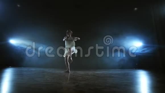 一个穿着白色连衣裙的年轻女孩在舞台上跳着现代芭蕾烟雾缭绕在蓝色的聚光灯下视频的预览图