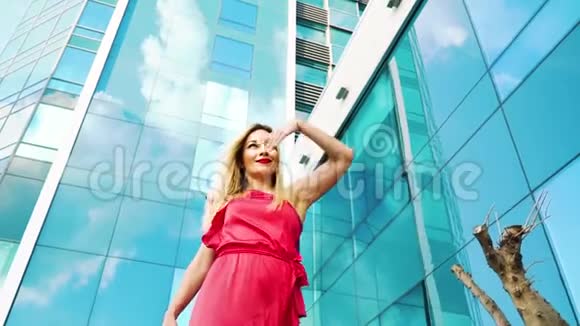 从摩天大楼经过的一位身穿红色裙子的金发美女低角度拍摄视频的预览图