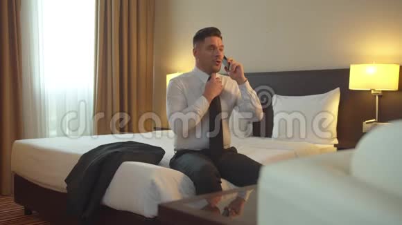 穿着衬衫和领带的人坐在酒店的床上打电话脱鞋视频的预览图