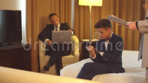 男人在笔记本电脑上工作小男孩坐在平板电脑上女人在酒店房间里拿着文件打电话视频的预览图