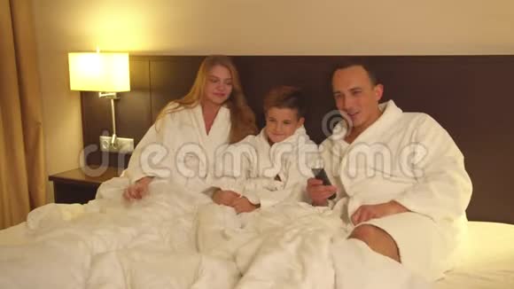 父母和小儿子坐在床上在旅馆房间里看电视视频的预览图