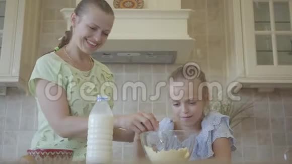 年轻的微笑的女人和她的小女儿在家里厨房里做蛋糕女孩在母亲面前拌面粉视频的预览图