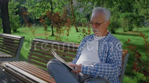 一个年长的男人坐在长凳上在公园里读一本有趣的书视频的预览图